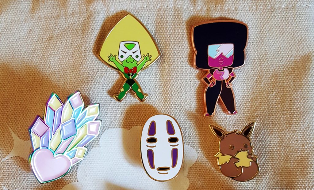 Starting an Art Business. Group of 5 enamel pins displayed on a canvas tote bag. Top row has two Steven Universe pins (Peridot and Garnet), while the second has a heart with crystals coming out of it, No Face from Spirited Away, and an Eevee (Pokemon).