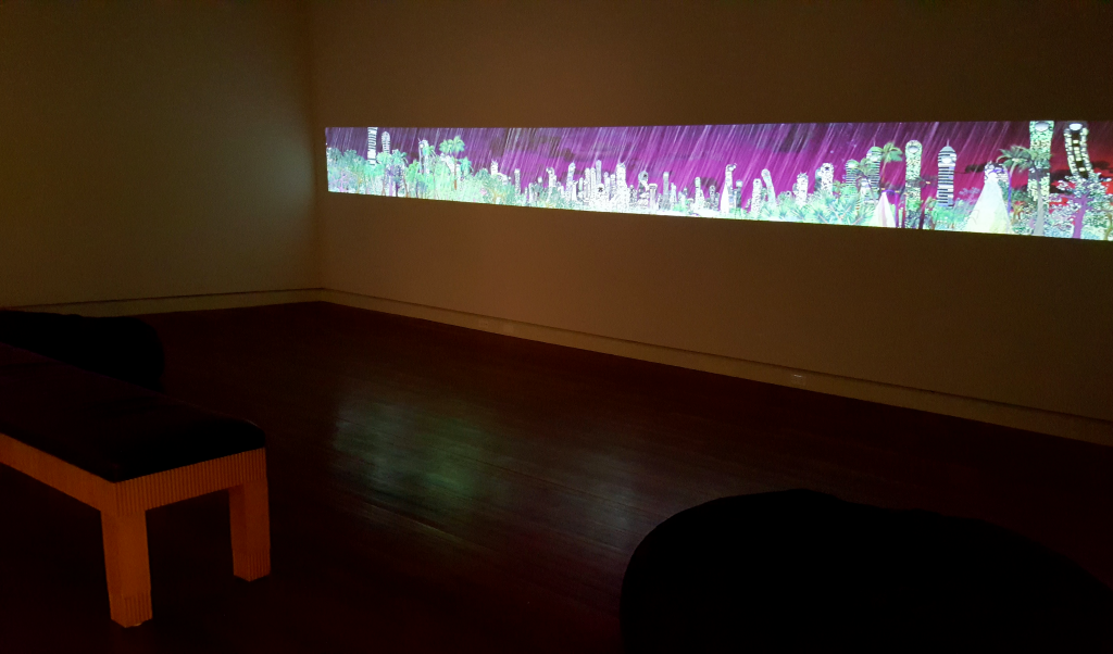 Chiho Aoshima City Glow Room with wide video art installation projected as a narrow rectangle along the right-hand wall. In this freeze-frame, there is a light colored skyline of plants and distant buildings, set against a dark purple sky. It is raining heavily. 