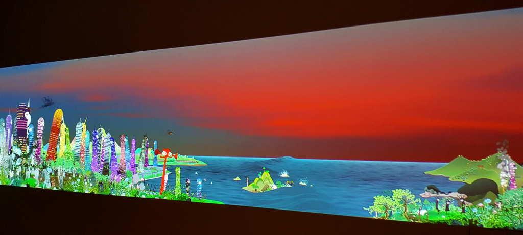 Chiho Aoshima Takaamonohara freeze-frame showing the video projection at an angle, focused on a cluster of anthropomorphic buildings on the far left end of the screen, all turned to watch something in the distance.