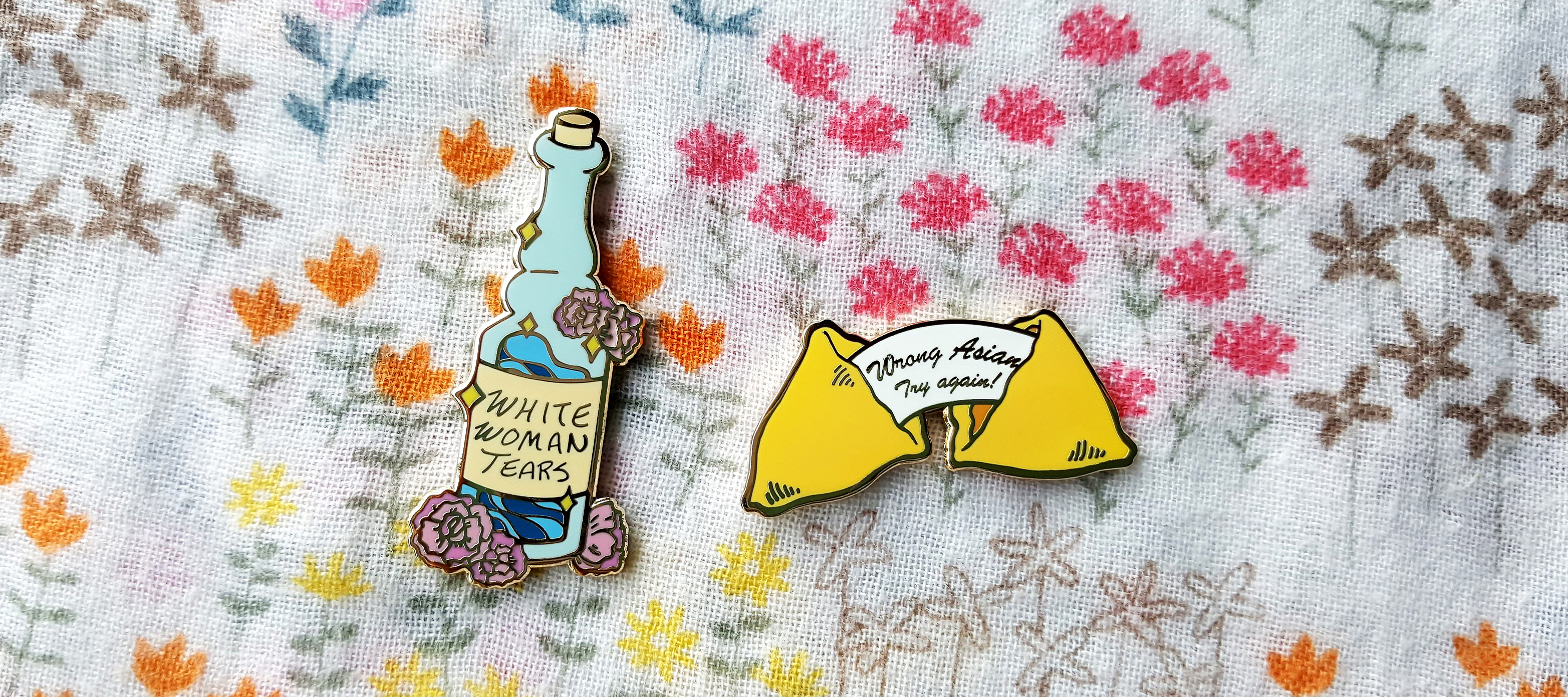 Two pins on a floral cloth background. The first is shaped like a bottle and reads "White Woman Tears." The second is a fortune cookie with the message "Wrong Asian Try Again!"